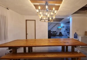 Comedor con mesa de madera y bancos, una araña y una puerta que da a otra habitación.