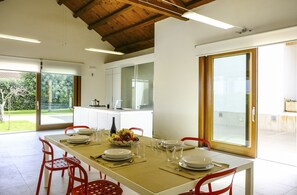 A dining area with a table set for four, a kitchen in the background, and a view of a garden through a glass door. - Villa in Sicily With Pool Near Erice & Trapani (Marsala)