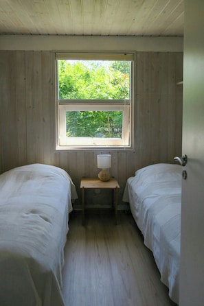 A room with two single beds, a small table with a lamp, and a window with a view of greenery. - Timber house with large garden 70 m from beach (Knebel)