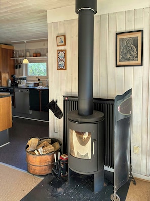 A kitchen with a wood-burning stove, a dining area, and a view of the outdoors. - Timber house with large garden 70 m from beach (Knebel)