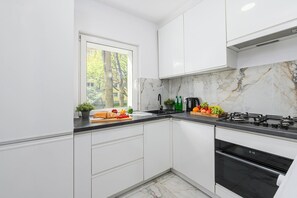 A modern kitchen with white cabinets, a black countertop, a gas stove, and a window with a view of trees. - Warsaw City Stay for Families (Warsaw)