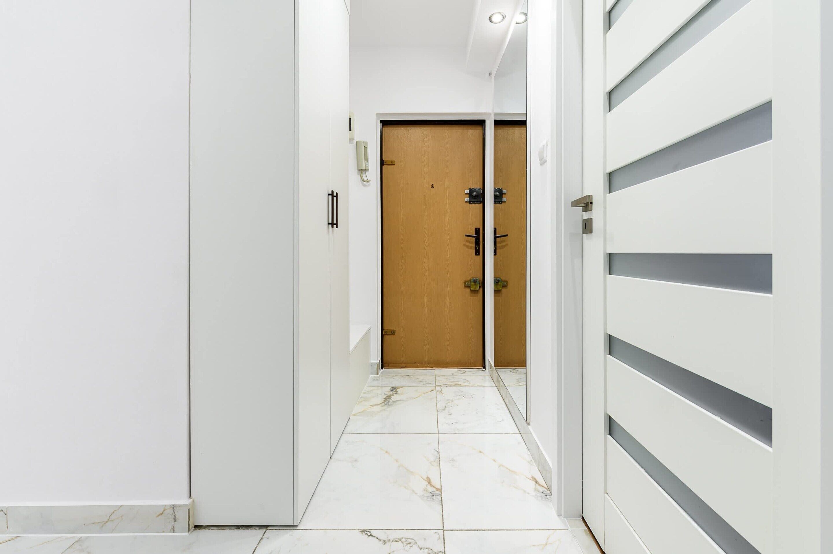A hallway with a wooden door, a white wall, and a white door with horizontal slats.