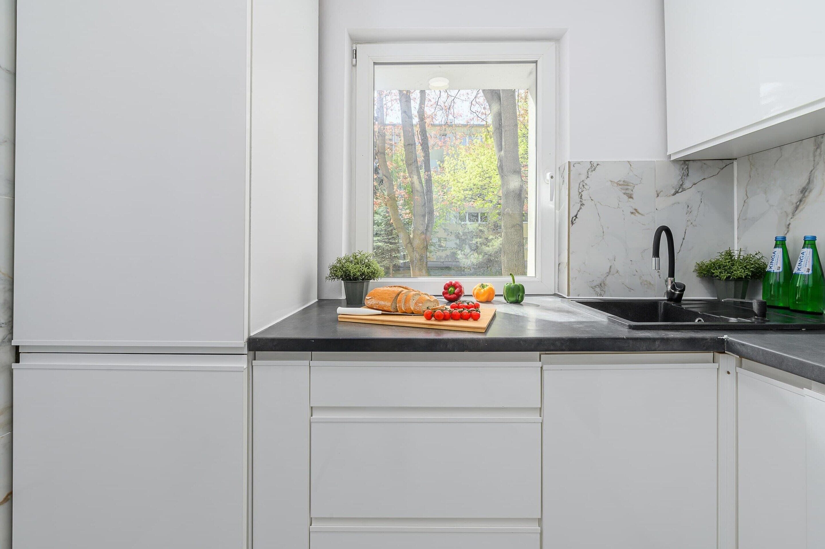 A kitchen with white cabinets, a marble backsplash, a window with a view of trees, and a countertop with a loaf of bread, tomatoes, and peppers.