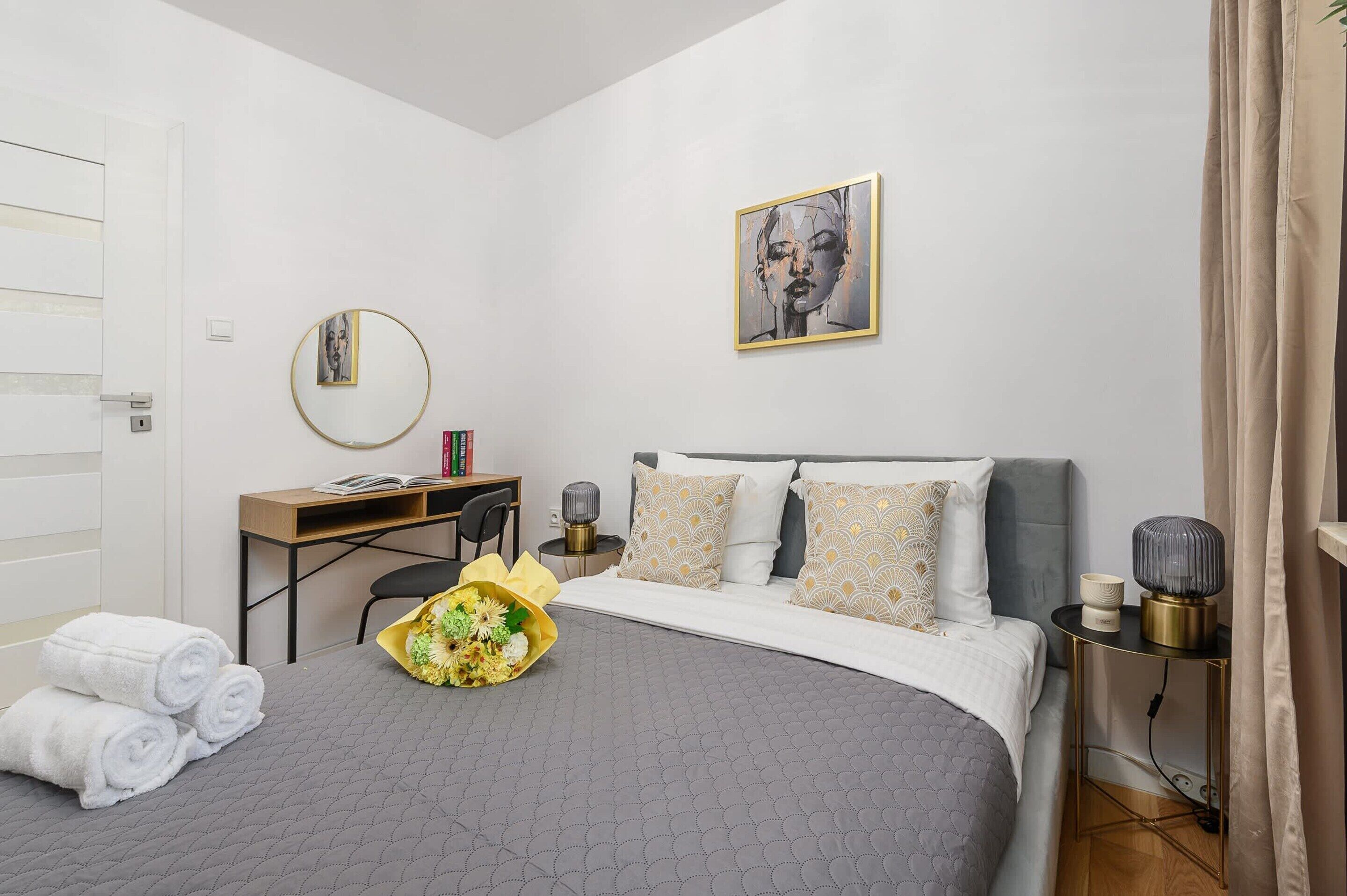 A hotel room with a bed, a desk, a chair, a round mirror, a bedside table with a lamp, and a wall with a framed picture.