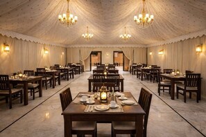 Un amplio salón de banquetes con varias mesas dispuestas para cenar, candelabros e iluminación proporcionada por apliques de pared.