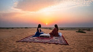 Two individuals sitting on a blanket on a sandy beach with a sunset in the background. - Marawi Desert Resort-Luxury Swiss Tents in Jaisalmer (Sam)