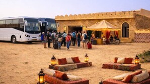 A desert setting with a caravan of buses parked in front of a building with a traditional design, surrounded by sand and outdoor seating with lanterns. - Marawi Desert Resort-Luxury Swiss Tents in Jaisalmer (Sam)