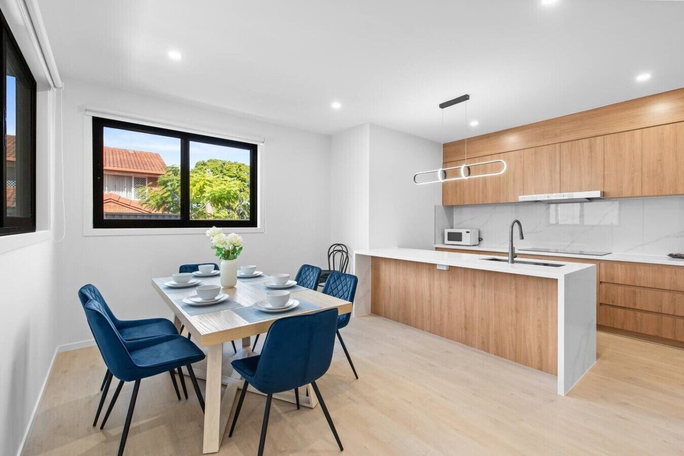 A modern kitchen with wooden cabinets, a white countertop, and a dining area with blue chairs and a table set for four.