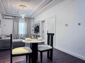 A modern living room with a sofa, a dining table with chairs, and a television mounted on the wall. - Stylish 1-Bedroom Apartment in Lekki 1 with Fast Wi-Fi & 24/7 Power (Lagos)