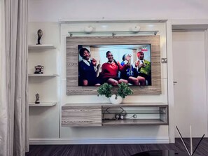 A flat-screen TV mounted on a wall with a wooden panel design, a small shelf with a decorative item, and a potted plant below the TV. - Stylish 1-Bedroom Apartment in Lekki 1 with Fast Wi-Fi & 24/7 Power (Lagos)