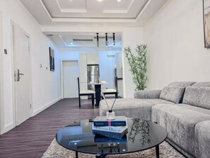 Ein modernes Wohnzimmer mit einer Couch, einem Couchtisch mit Büchern, einem Essbereich mit Stühlen und einer Küche im Hintergrund.