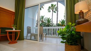 A room with a sliding glass door leading to a balcony with a white railing, a wicker chair, a small table with a plant, and a lamp.