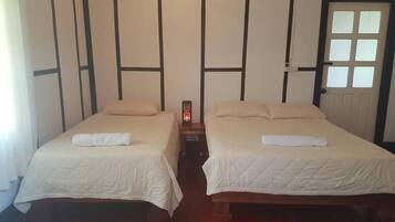 A room with two beds, each with a white bedspread and a pillow, a small table with a decorative item, and a door with a window.