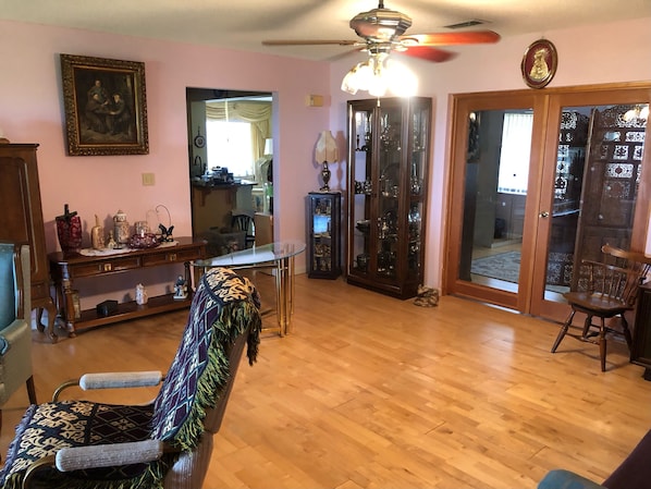 A living room with a wooden floor, a patterned armchair, a glass-top coffee table, a ceiling fan, and a door leading to a balcony.