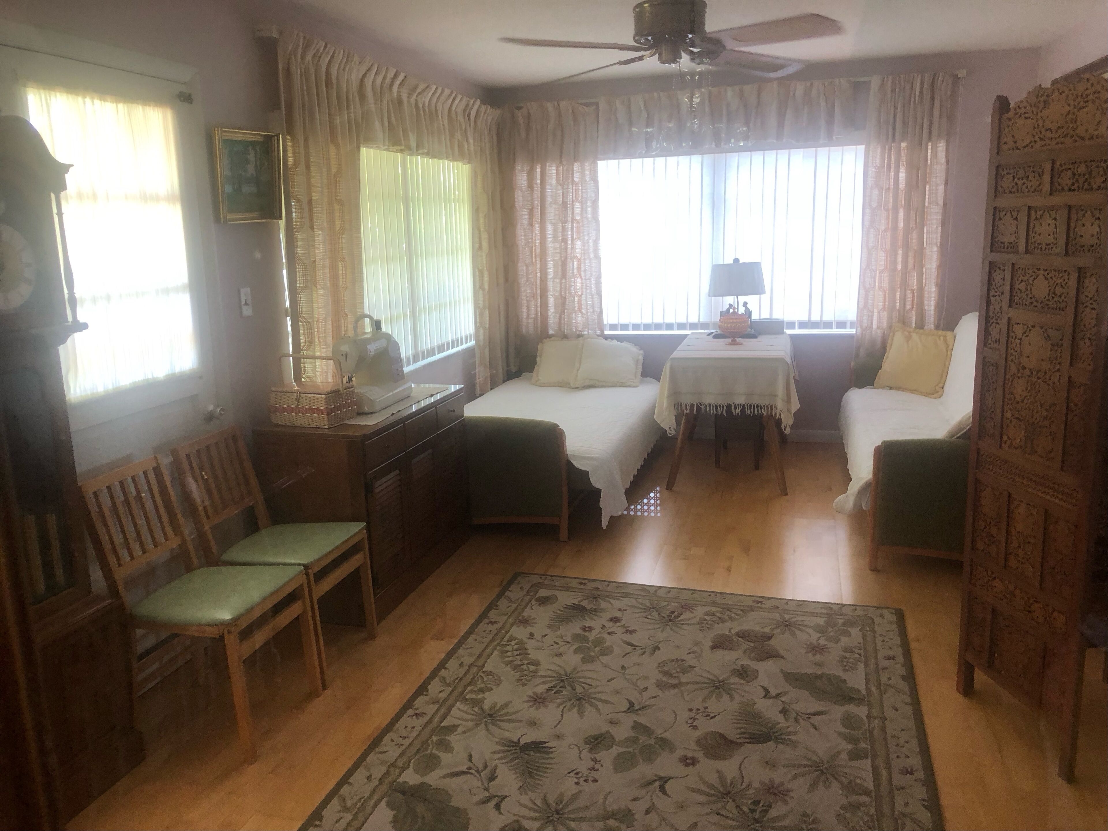 A room with a bed, a desk with a sewing machine, a chair, a small table with a lamp, and a decorative wall panel.