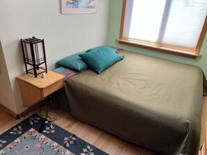 Un dormitorio con cama, mesita de noche, lámpara, ventana con persianas y una alfombra estampada.