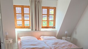 Ein Schlafzimmer mit Holzboden, einem Bett mit weißem Gestell, zwei Fenstern mit Vorhängen und einem Nachttisch mit Lampe.