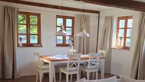 Ein Zimmer mit einer Couch, einem Esstisch, Stühlen und fensterbelaubten Fenstern.