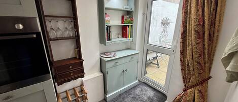 Una cocina compacta con horno integrado, copas de vino en una estantería y una ventana pequeña con cortinas.