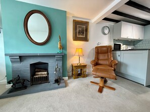 A living room with a fireplace, a large mirror, a brown leather chair, a small table with a lamp, and a kitchen area with cabinets and appliances.