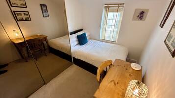 Un dormitorio con cama, escritorio, silla, ventana con persianas y mesita de noche con lámpara.