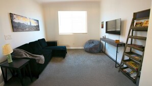 A living room with a dark-colored sofa, a small round ottoman, a lamp, a bookshelf, and a painting on the wall. - Star Light, Estrella Mountain Ranch (Goodyear)