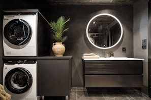 A modern bathroom with a washing machine, a potted plant, a round mirror, and a sink with towels.