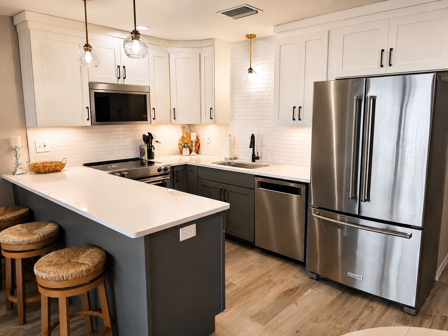 Una cocina moderna con un refrigerador de acero inoxidable, un microondas y un lavavajillas. Hay taburetes de madera y una isla grande con encimera blanca.