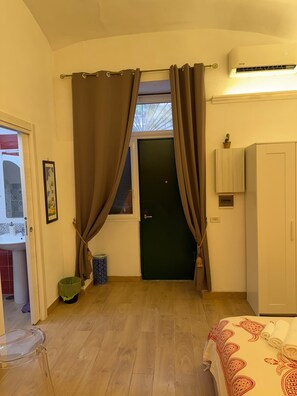 A room with a wooden floor, a door with curtains, a small trash bin, a framed picture, and a bed with a patterned blanket. - Luchino's Den (Napoli)