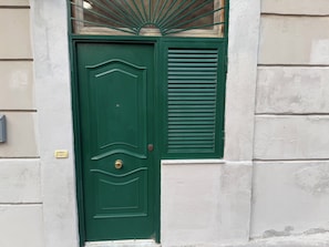 A green door with a brass handle and a window with a sunburst design above it. - Luchino's Den (Napoli)