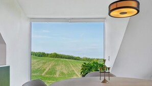 A dining area with a view of a green field through a large window, a round table, and a hanging light fixture. - 4 Star Holiday Home in Allinge (Allinge)