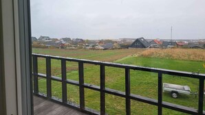 A view from a balcony with a wooden railing, overlooking a residential area with houses and a grassy field. - 4 Star Holiday Home in Allinge (Allinge)