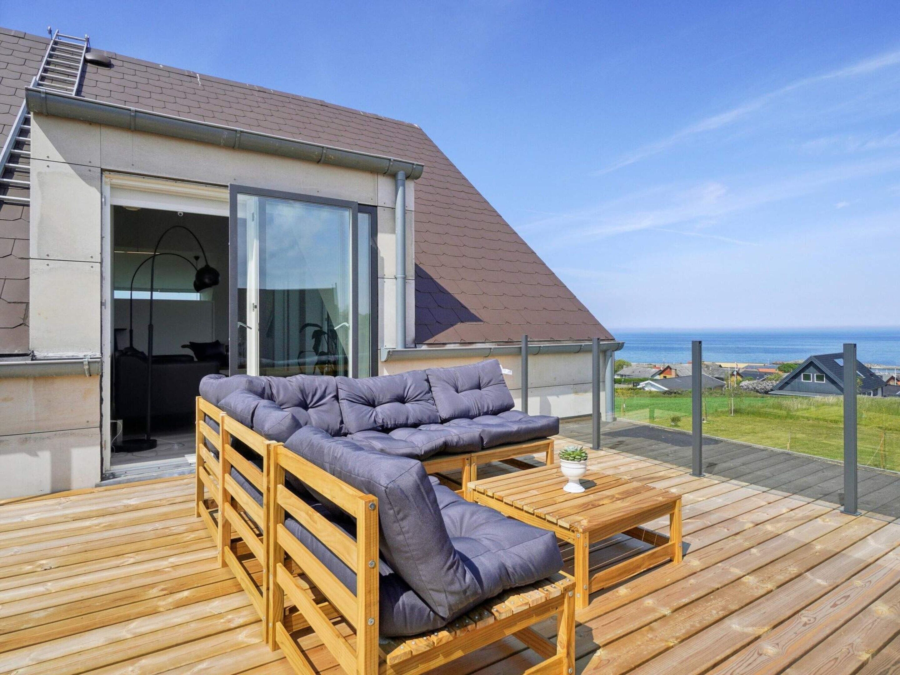 En træterrasse med en sofa og et bord, en glasdør der fører indendørs, og udsigt til havet.