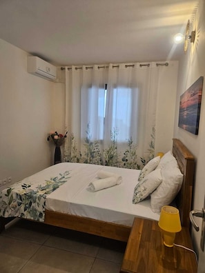 A hotel room with a bed, a bedside table with a lamp, a window with curtains, an air conditioning unit, and a wall-mounted picture. - Un T2 récent  , confortable  dans un petit  immeuble au coeur de Saint Denis (Saint-Denis)