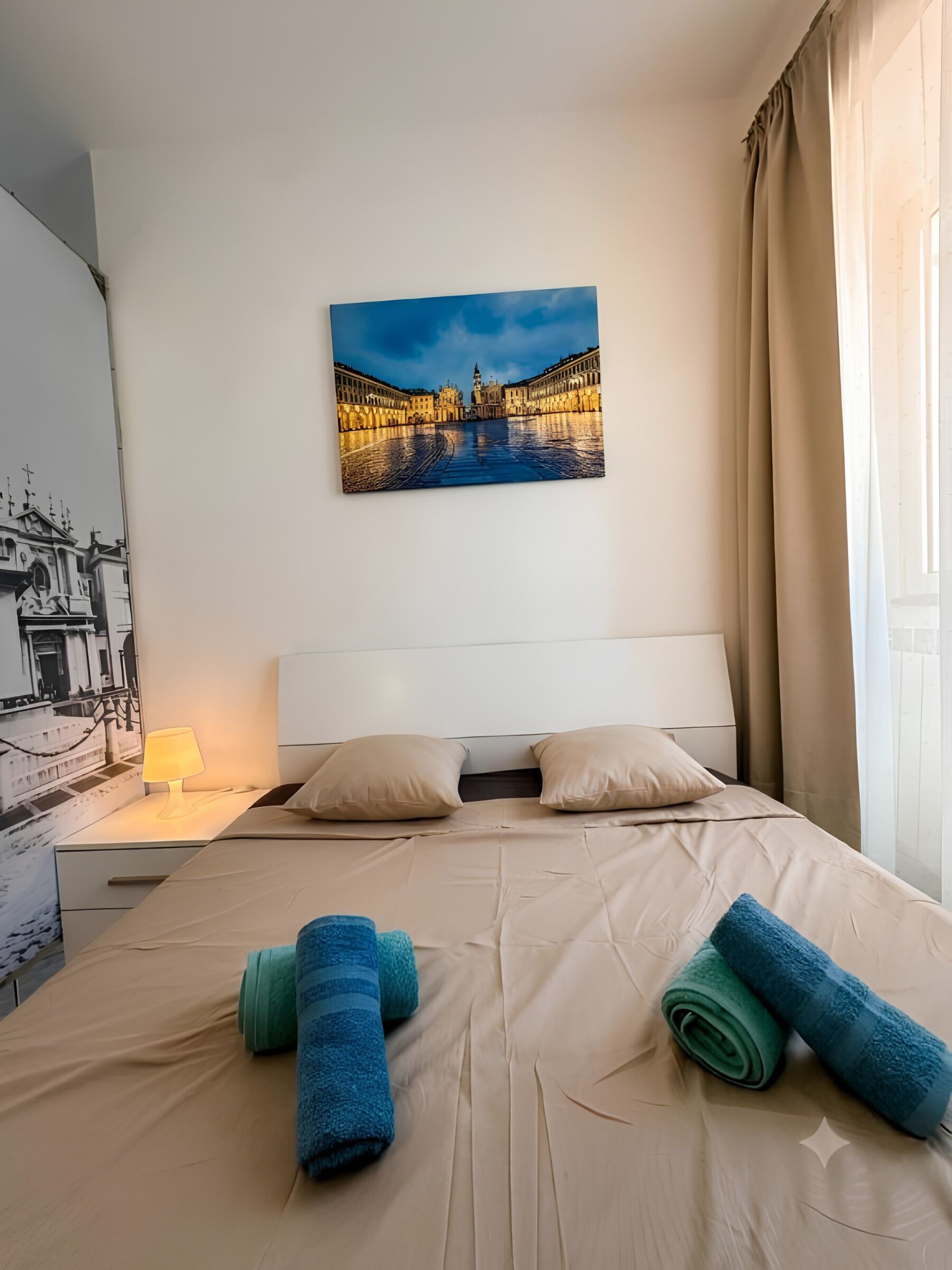 Una camera d'albergo con un letto, cuscini, un comodino con una lampada, un quadro appeso al muro e una finestra con tende.