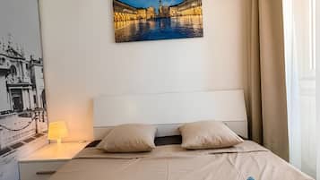 Una camera d'albergo con un letto, cuscini, un comodino con una lampada, un quadro appeso al muro e una finestra con tende.