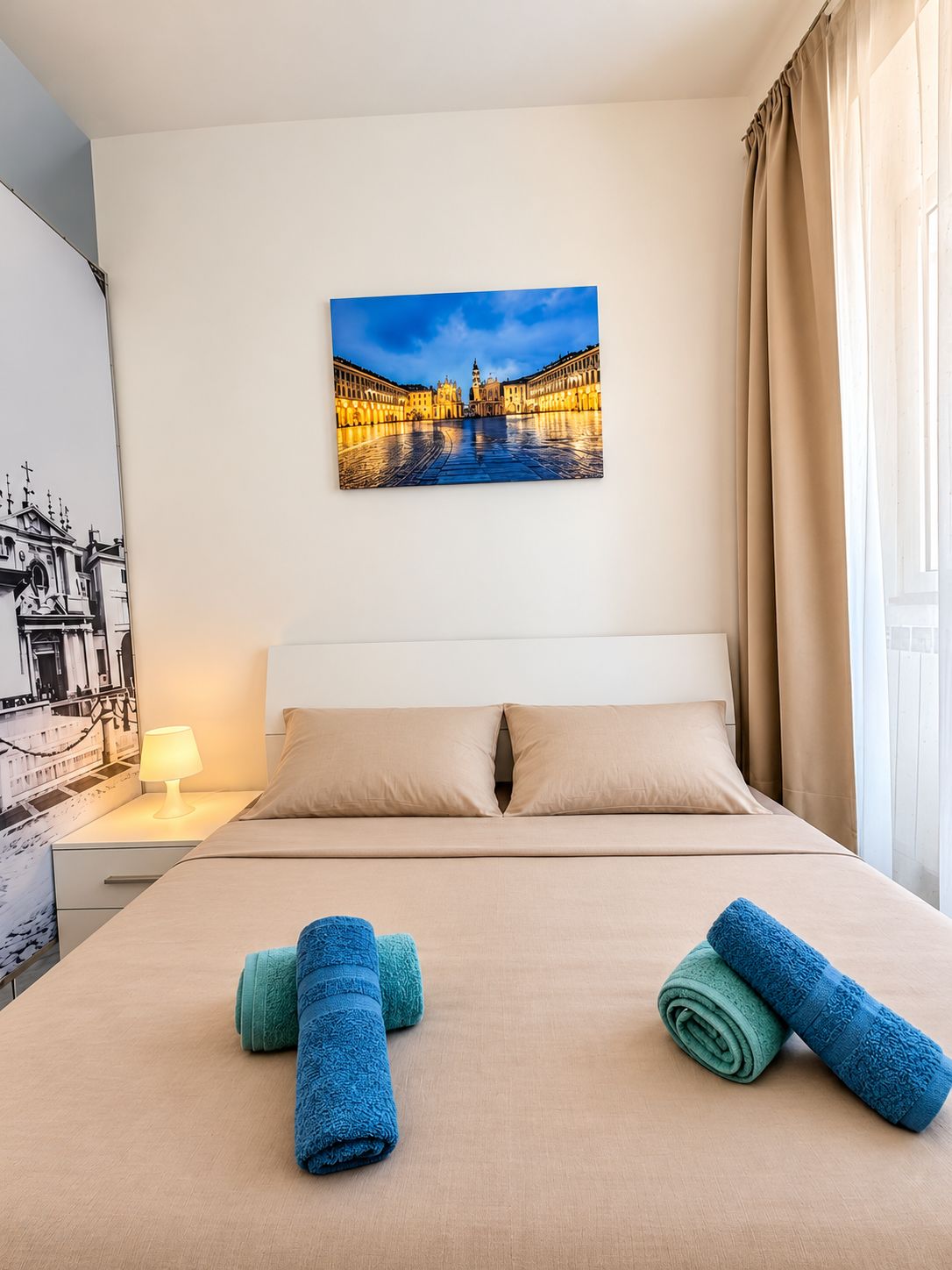 Une chambre d’hôtel avec un lit, deux oreillers, une table de chevet avec une lampe et un tableau représentant un paysage urbain accroché au mur.
