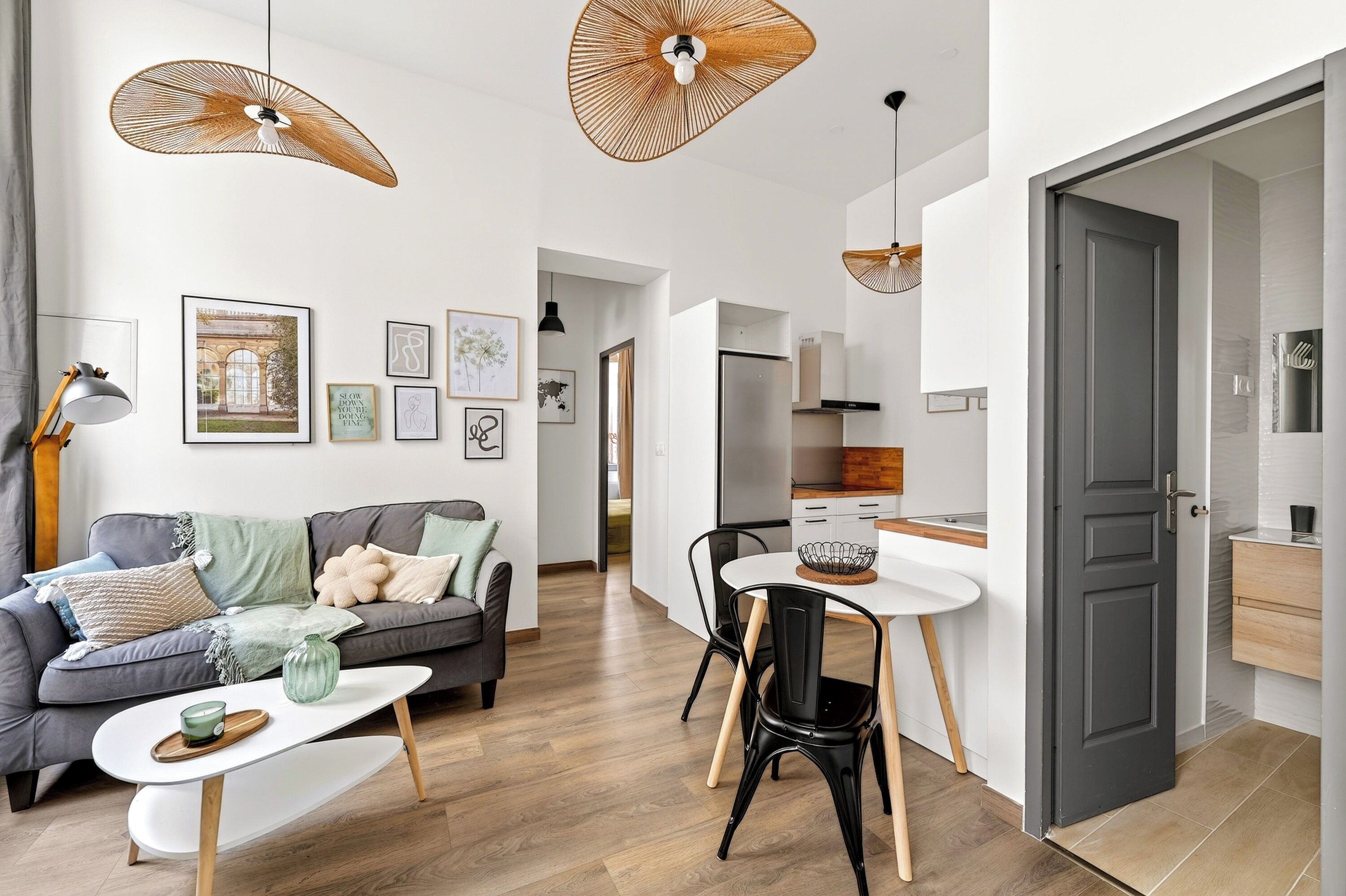 Un salon moderne avec un canapé gris, une table basse et un coin repas comprenant une table et des chaises blanches. Deux lampes suspendues sont fixées au plafond.
