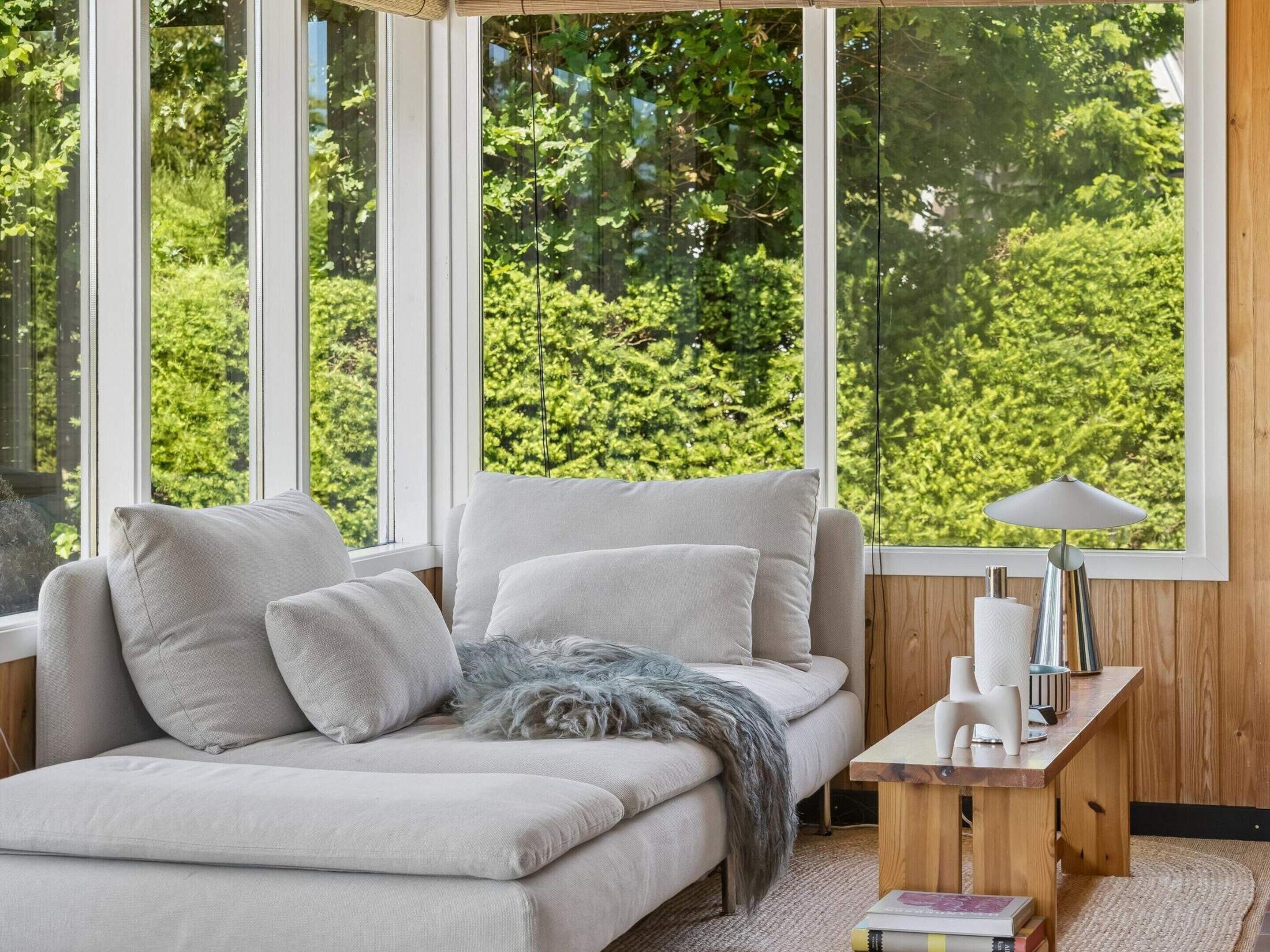 A modern living room with a sofa, a lamp, and a view of greenery through large windows.