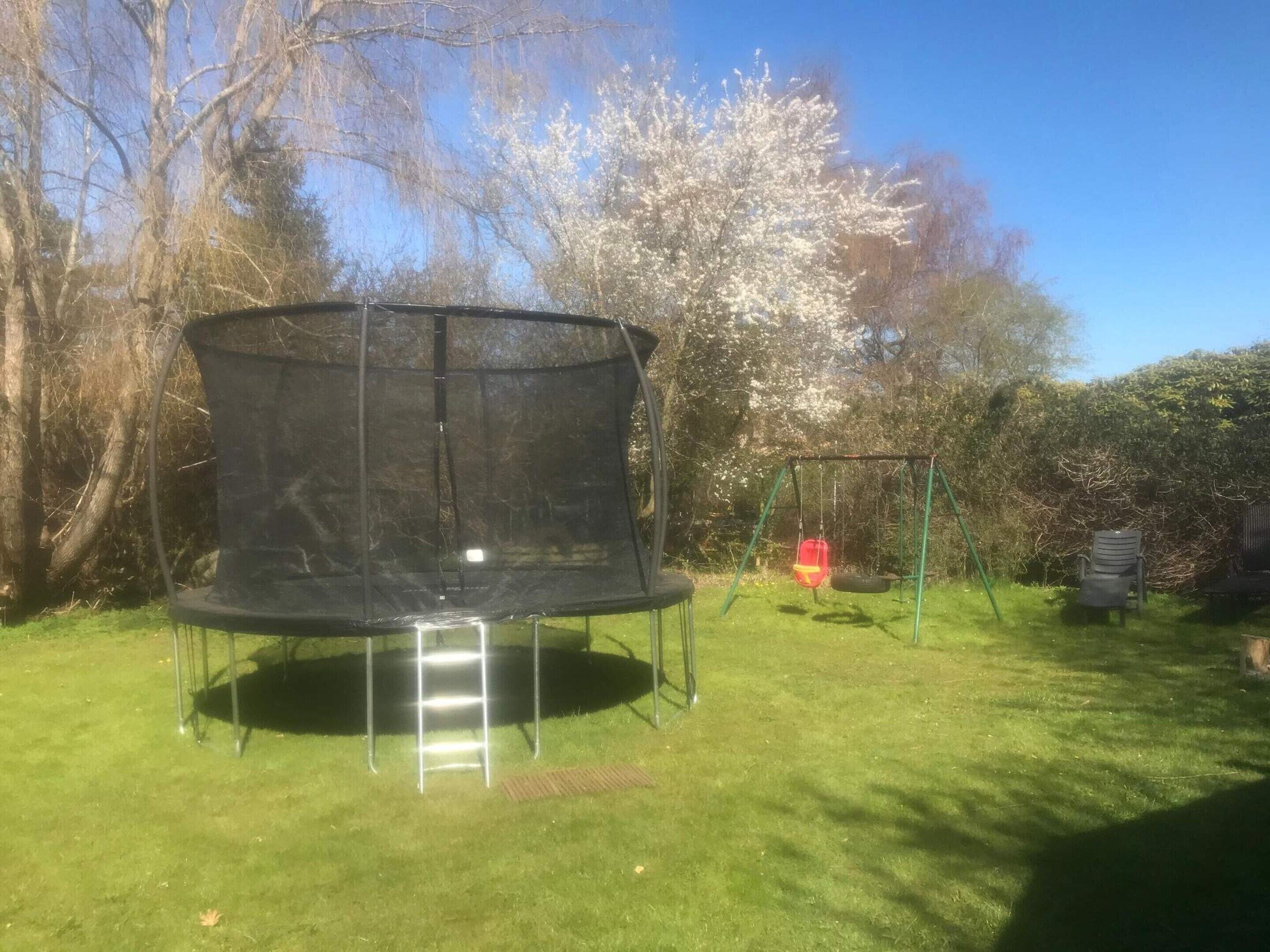 A trampoline with a safety net, a swing set, and a bench in a grassy yard with trees in the background.
