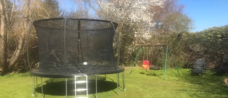 A trampoline with a safety net, a swing set, and a bench in a grassy yard with trees in the background.