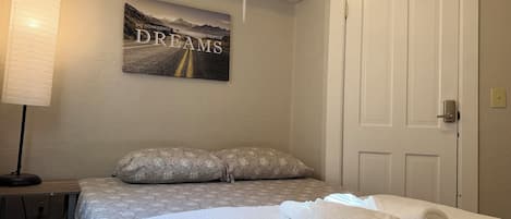 Una cama bien hecha con un cojín, una lámpara de noche y un cartel motivacional en la pared.