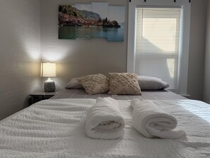 A hotel room with a bed, a nightstand, a lamp, a painting of a coastal scene, and a window with blinds. - Cozy Home close to Notre Dame & Downtown Southbend (South Bend)
