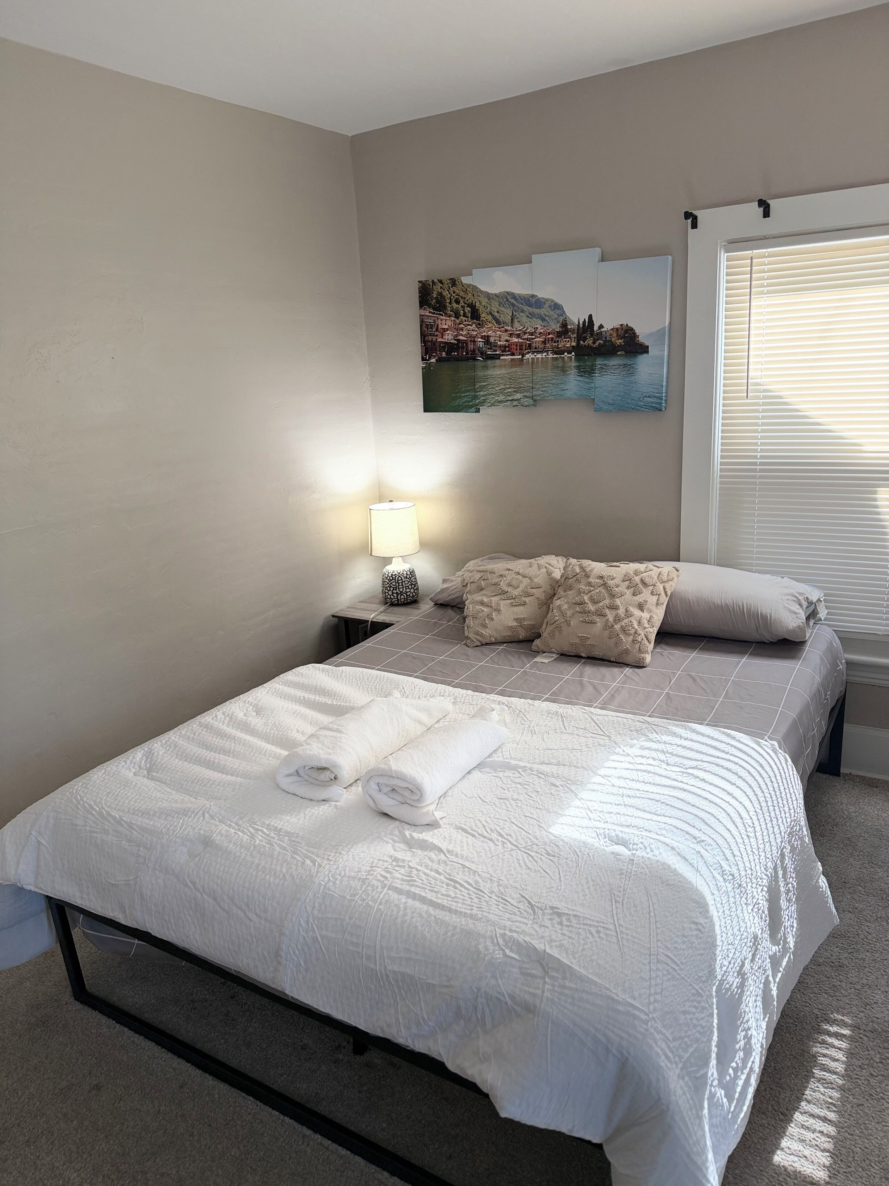 Una cama bien hecha con sábanas blancas y toallas dobladas, una lámpara de noche y un cuadro con una escena costera en la pared.
