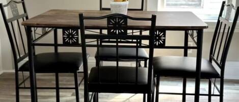 A dining table with four black chairs, a white vase with yellow flowers, and a window in the background.