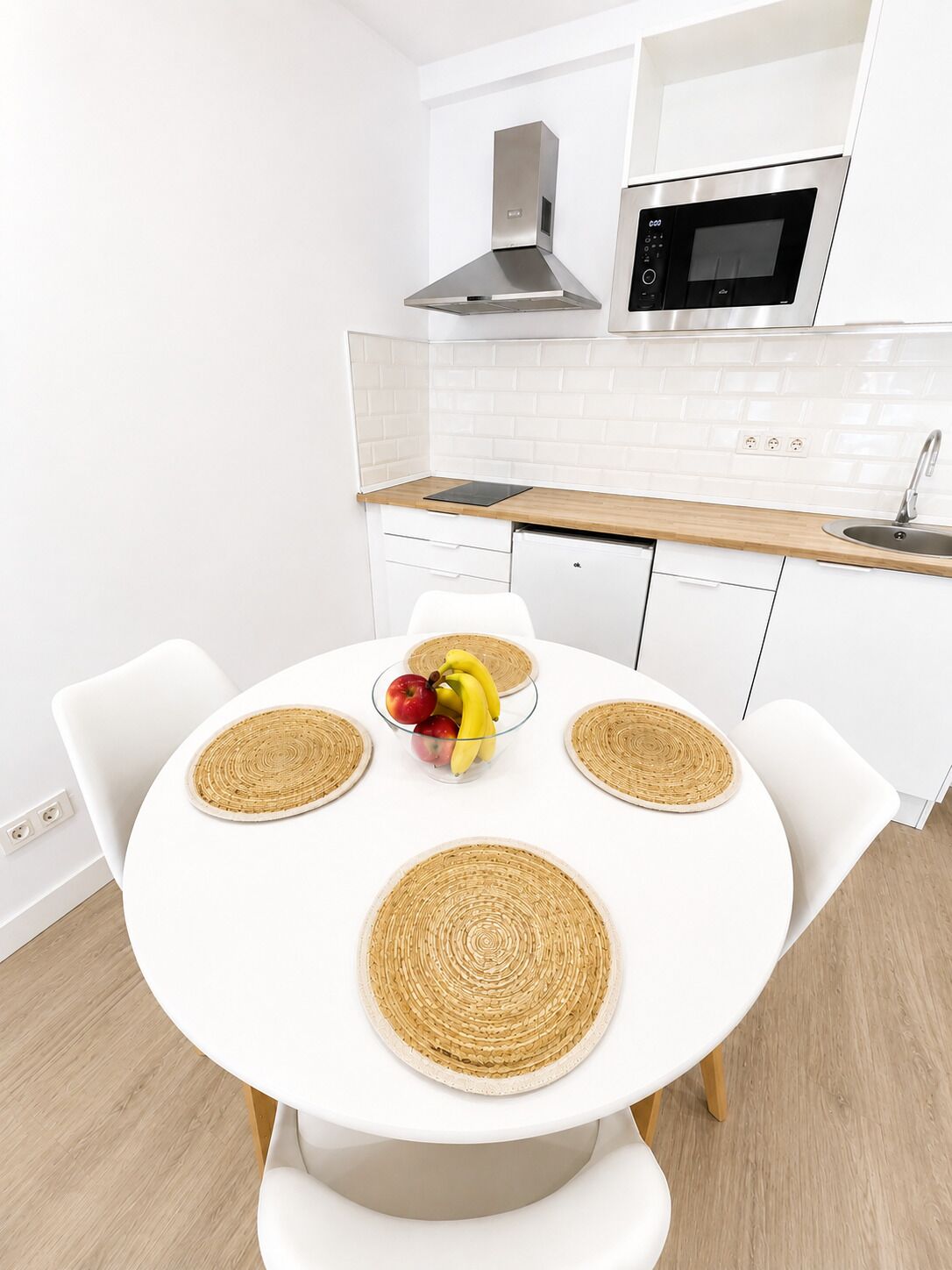 A modern kitchen with white cabinetry, a stainless steel range hood, a microwave, and a round dining table with woven placemats and a fruit bowl.