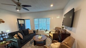 Ein Wohnzimmer mit einem dunkelgrauen Sofa, einem runden Couchtisch, einem braunen Ledersessel, einem flachbildfernseher an der Wand und einem Deckenventilator.