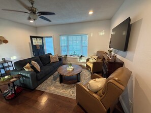 Ein Wohnzimmer mit einem dunkelgrauen Sofa, einem runden Couchtisch, einem braunen Ledersessel, einem flachbildfernseher an der Wand und einem Deckenventilator.