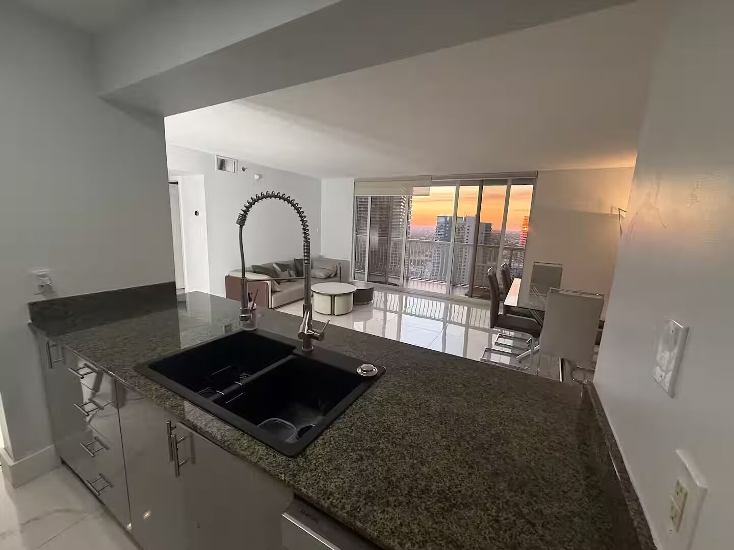 A modern kitchen with a built-in sink, a dining area with a round table, and a view of a cityscape through large windows.