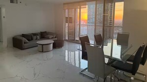 A modern living room with a sofa, a round coffee table, and a glass dining table with chairs. Large windows provide a view of the cityscape during sunset. - Brickell Penthouse Breathtaking Views (Miami)